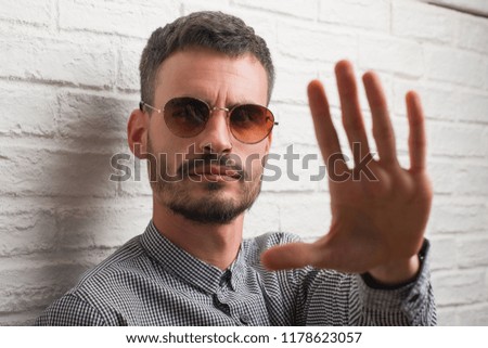 Young adult man wearing sunglasses standing over white brick wall with open hand doing stop sign with serious and confident expression, defense gesture