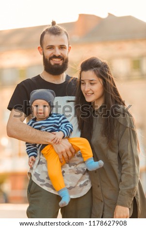Happy young family. Stylish bearded man and young beautiful wife with a little child in their arms, hugging, kissing and playing walking around the city.