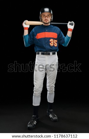 Full length portrait of a baseball player posing with a bat on black background. Young American sportsman in a uniform.