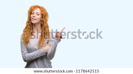 Young redhead woman with a big smile on face, pointing with hand and finger to the side looking at the camera.