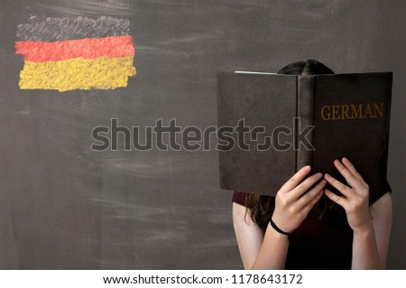 A young girl reading and studying a German text book sitting in front of a chalkboard with a German flag drawn in coloured chalk in the top left corner of the black board.