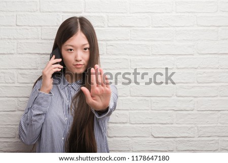 Young Chinese woman over brick wall talking on the phone with open hand doing stop sign with serious and confident expression, defense gesture