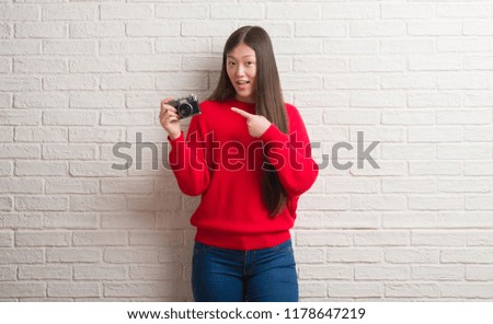 Young Chinese woman over brick wall holding vintage camera very happy pointing with hand and finger