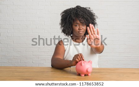 Young african american woman sitting on the table holding piggy bank with open hand doing stop sign with serious and confident expression, defense gesture