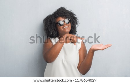 Young african american plus size woman over grey grunge wall wearing fashion sunglasses amazed and smiling to the camera while presenting with hand and pointing with finger.