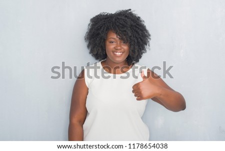 Young african american woman over grey grunge wall doing happy thumbs up gesture with hand. Approving expression looking at the camera with showing success.