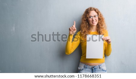 Young redhead woman over grey grunge wall holding blank paper sheet surprised with an idea or question pointing finger with happy face, number one