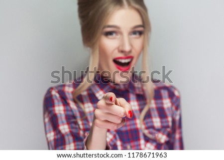 Amazed beautiful blonde girl with pink checkered shirt, collected hairstyle and makeup standing, looking at camera and with finger pointing and shocked. indoor studio shot, isolated on gray background