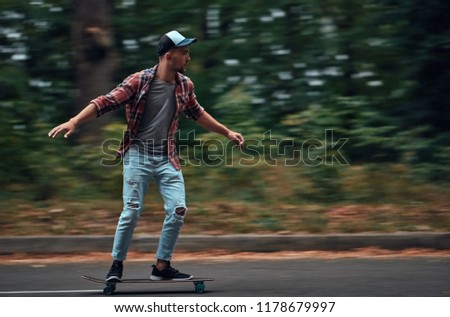 A stylish hipster man in a red shirt and a baseball cap rides at high speed on a skateboard on the road in the middle of the forest.