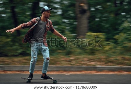 A stylish hipster man in a red shirt and a baseball cap rides at high speed on a skateboard on the road in the middle of the forest.