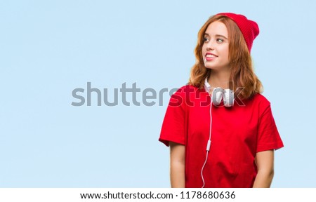 Young beautiful hipster woman over isolated background wearing headphones and cap looking away to side with smile on face, natural expression. Laughing confident.