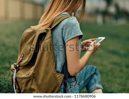 A hipster girl with a vintage backpack holds a smart gadget. A traveler using a mobile phone in a woman's hand and sitting on the grass.