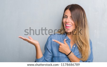 Young adult woman over grunge grey wall wearing denim outfit amazed and smiling to the camera while presenting with hand and pointing with finger.