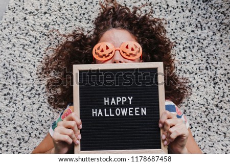 young woman lying on the floor wearing orange halloween glasses and holding a letter board with happy halloween message. Concept, lifestyle indoors