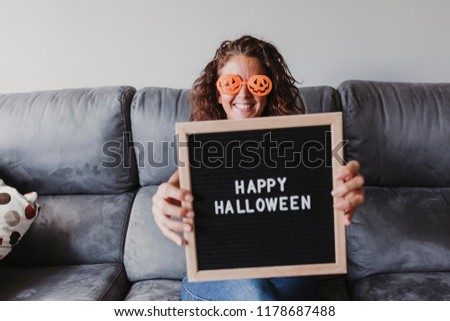 young woman sitting on the sofa, smiling and wearing orange halloween glasses. holding a letter board with happy halloween message. Concept, lifestyle indoors