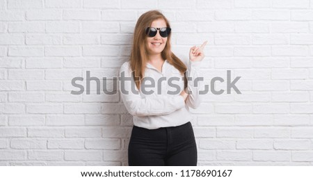 Young adult business woman standing over white brick wall wearing sunglasses very happy pointing with hand and finger to the side