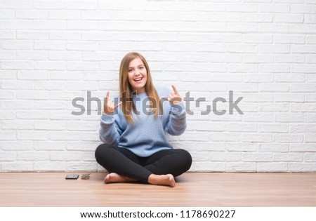 Young adult woman sitting on the floor in autumn over white brick wall shouting with crazy expression doing rock symbol with hands up. Music star. Heavy concept.