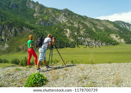 Young man photographer and red haired girl travels through the Altai with the camera on the tripod takes a shot of the snow-covered mountains and the rocks with turquoise winding river Katun
