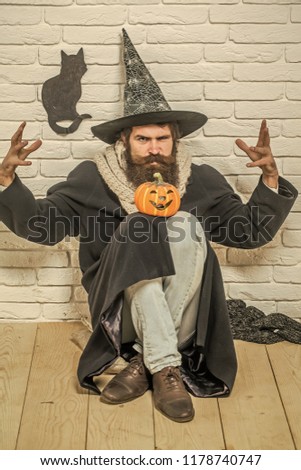 Halloween evil spell. Man in witch hat doing magic on floor. Bad luck concept. Hipster with pumpkin and black cat symbol on wall. Autumn holidays celebration.