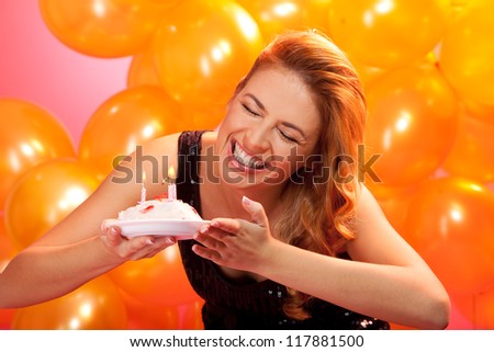closeup portrait of joyful young woman holding birthday cake with candles against balloons