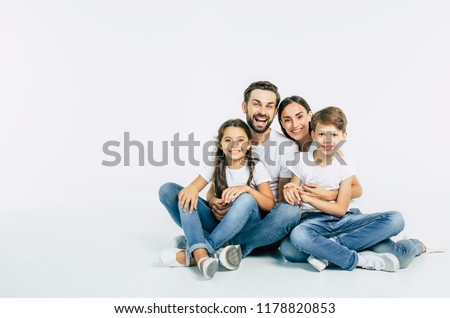 Cool team. Beautiful and happy smiling young family in white T-shirts are hugging and have a fun time together while sitting on the floor and looking on camera.