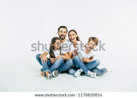 Father, mother, son and cute daughter. Beautiful and happy smiling young family in white T-shirts are hugging and have a fun time together while sitting on the floor and looking on camera.