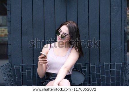 Portrait of young charming woman texting using mobile phone. Pretty dark-haired girl sitting outside in sunglasses. Technology and communication tool concept