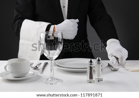 Closeup of a waiter in a tuxedo setting a formal dinner table. Shallow depth of field in horizontal format on a light to dark gray background. Man is unrecognizable. Royalty-Free Stock Photo #117891871