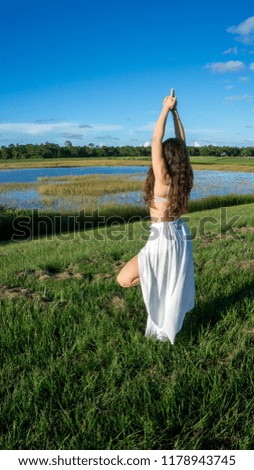 Young brunette spanish woman doing Vriksasana tree yoga pose in a field next to a lake with long curly hair Space in sky