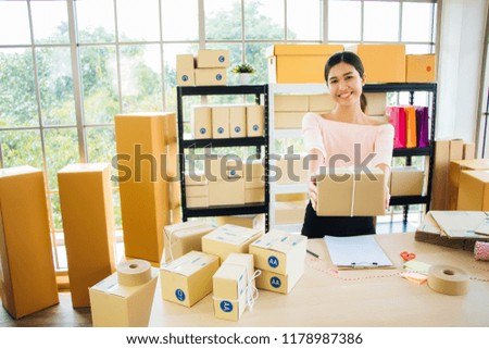 Asian girl in pink casual dress Smiling and holding the finished goods, handed out to prepare the goods to send to customers. In the office filled with product boxes. And packing equipment. Royalty-Free Stock Photo #1178987386