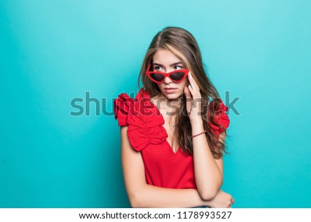 Elegant young woman wearing summer dress and sunglasses, thinking about her summer vacation. Woman with hand on chin, isolated over pastel blue background.