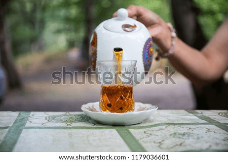 Eastern black tea in glass on a table in forest. Hand pouring tea in glass. Armudu traditional cup. Green nature background. Selective focus