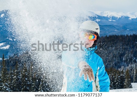 a woman in a blue ski suit and mask hurls snow at the camera on top of the mountain.the girl throws snow at the camera
