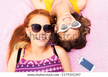 summer fashion, leisure and technology concept - smiling teenage girls in sunglasses and earphones lying on picnic blanket and listening to music from smartphone