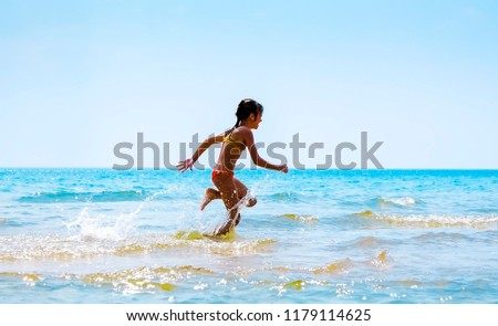 Picture of exciting little girl running on beach beside waves. Vintage image of kid in day sunlights on seaside background.
