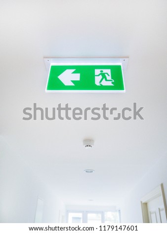 Emergency Exit sign (left arrow) and Smoke sensor hang down on ceiling at the bright white interior corridor with cream colored wooden door on the right hand side