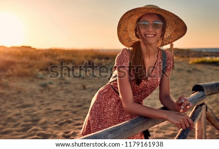 Beautiful smiling girl in sunglasses and summer hat overlooking the sunset landscape on the beach and leaning against wooden fence.