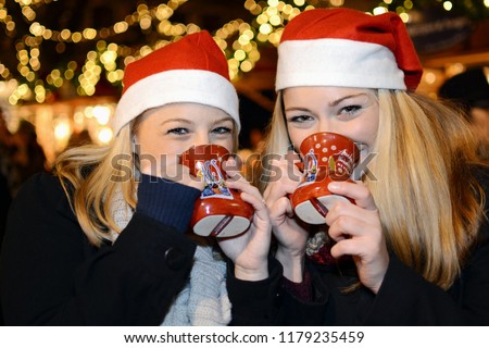 Girlfriends visit Christmas market with many stalls and drink mulled wine from cups with the inscription "Aachener Weihnachtsmarkt" which means Aachen Christmas market