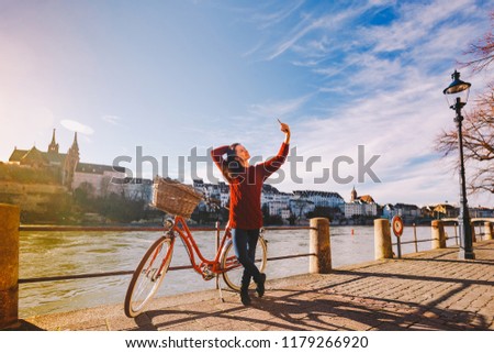 A beautiful young woman with a retro red bicycle is making a photo of herself in the old city of Europe on the River Rhine embankment in the Swiss city of Basel. Sunny warm day in winter.