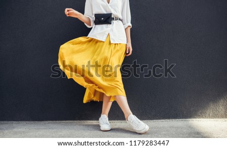 Horizontal cropped image of stylish slim woman in beautiful yellow skirt. Caucasian female fashion model standing over gray wall background outdoor with copy space. Royalty-Free Stock Photo #1179283447
