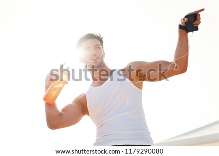 Athletic muscular man feeling victory and enjoys success holding a bottle of water. Emotion, power, gesture - young man celebrating victory with sun light in background.
