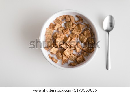 Cinnamon cereal within a white bowl with a silver spoon go the right of it.table top view, top layout and a close up of the details of the cereal.perfect white table for comfortable sit down breakfast