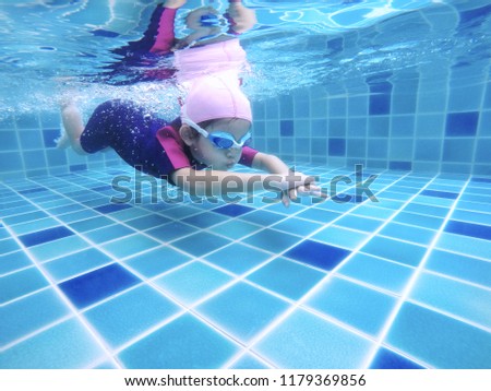 Underwater young little cute girl is swimming in the swimming pool with her swimming teacher. Seen under water while she is diving forward ahead of her. Swimming kid in the pool concept.