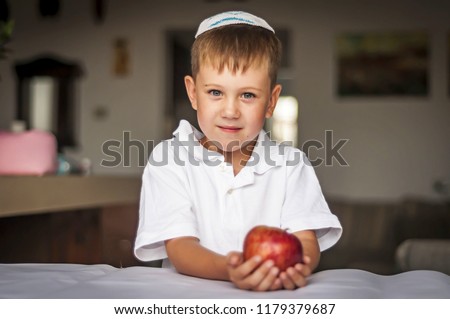 Cute Caucasian blue-eyed boy in a traditional Jewish white kippah cap with an apple in his hands. Russian Jewish boy celebrating Jewish New Year - Rosh Hashanah. Royalty-Free Stock Photo #1179379687