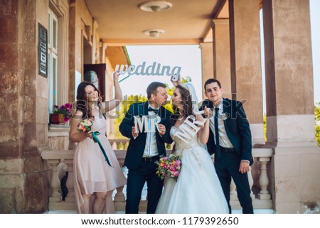 Newlywed couple, bridesmaids & groomsmen having fun outdoors before wedding ceremony. Bride and groom and friends hold in hand wooden wedding decoration with words "Wedding"