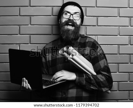 Bearded man, long beard. Brutal caucasian smiling happy unshaven hipster holding laptop and craft paper rolls in red black checkered shirt with hat and glasses on brown brick wall studio background