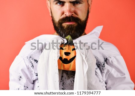 Man with strict face expression on red background. Guy with beard holds orange pumpkin with smile. Halloween and happy holiday concept. Halloween character in white long sleeved ghost costume