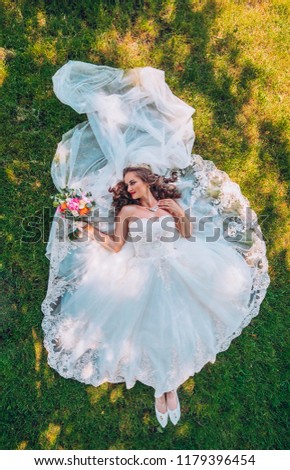 Beautiful, bride in a wedding dress lying at the grass. Wedding day. Bride posing on a green lawn. Long hair, sweet and gentle smile. Top view picture.Woman with long veil.