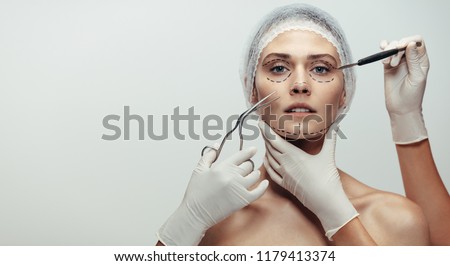 Horizontal shot of woman under going a face lift surgery. Female face with dotted line drawn and beautician holding surgical instruments on grey background. Royalty-Free Stock Photo #1179413374