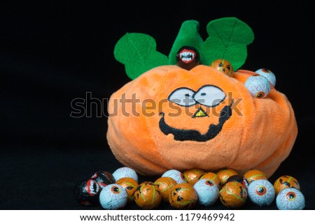 Funny plush toy in the shape of a halloween pumpkin with lots of chocolate balls around it, covered with nice smiley faces, on a black background. Useful to represent a funny event for halloween